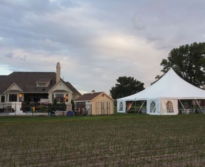 A white high-peak frame tent set up in a field of young crops next to a large house and shed under a dusky, cloud-filled sky.