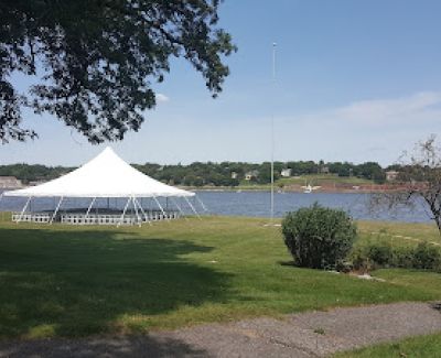 A white high-peak frame tent pitched on a grassy area overlooking a calm body of water under a clear blue sky.