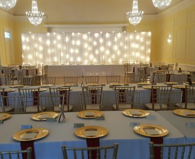 An elegantly decorated banquet hall with tables set with light blue cloths, gold charger plates, and gold Chiavari chairs, facing a head table backed by a wall illuminated with vertical string lights under chandeliers.