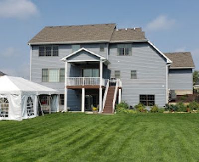 A residential backyard with striped mowing patterns on the lawn, featuring a white frame tent with windowed sidewalls set up near the back of a blue house.