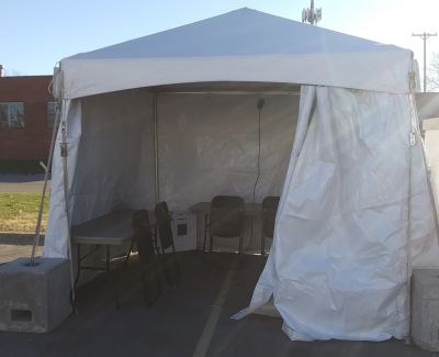 A small, square white frame tent set up in a paved parking lot, open at the front showing a table and chairs inside, secured with concrete block weights.