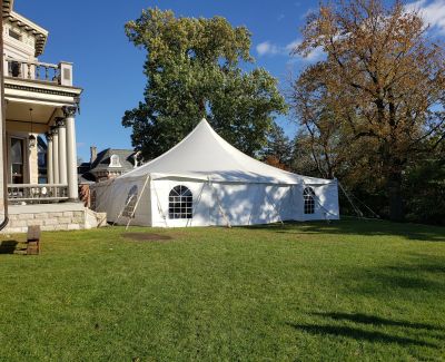 A white high-peak pole tent with arched window sidewalls pitched on the green lawn adjacent to a large, stately historic house on a sunny day.