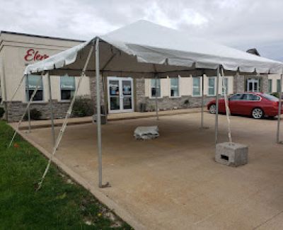 A small white frame tent set up on a paved area outside a commercial building entrance, secured by concrete block weights at the legs.
