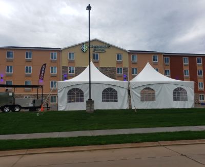 Two adjacent white high-peak frame tents with arched window sidewalls installed on a grassy area in front of a large, modern hotel building under an overcast sky.