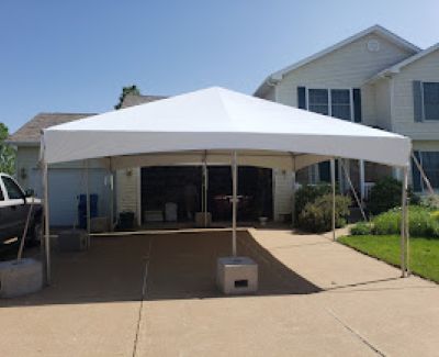 A white frame tent erected on a concrete residential driveway, positioned directly in front of the open garage door and secured with concrete weights.