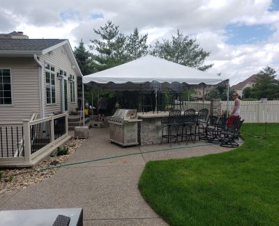 A white frame tent covering a backyard patio equipped with an outdoor kitchen island and bar stools, adjacent to the house and lawn.