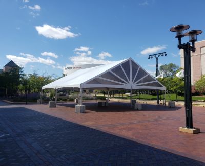 A white frame tent with a clear canopy section installed on a brick plaza in a public square or campus area, secured with concrete weights under a blue sky with white clouds.