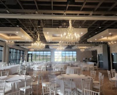 Interior of a spacious, modern banquet hall featuring large windows, coffered ceilings with chandeliers, and numerous round tables set with white linens and white chairs.