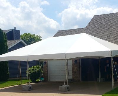 A white frame tent pitched on a concrete residential driveway in front of a house's garage, secured with concrete block weights under a partly cloudy sky.