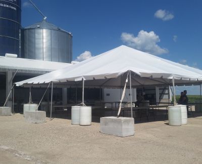 A large white frame tent erected on a gravel area next to large metal farm silos under a bright blue sky, with tables set up underneath and weights securing the legs.
