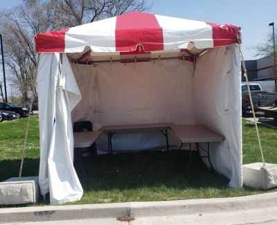A small, square white frame tent with a red-and-white striped valance, set up on grass near pavement with sidewalls partially open, revealing tables inside. It's secured with concrete weights.