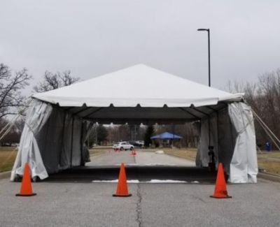 A white frame tent set up as a drive-through checkpoint or entrance on a paved road, marked by three orange traffic cones, with partially attached sidewalls.
