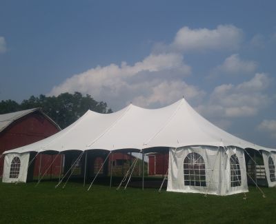 A large white high-peak pole tent with windowed sidewalls pitched on a green lawn in front of a red barn, under a partly cloudy blue sky.