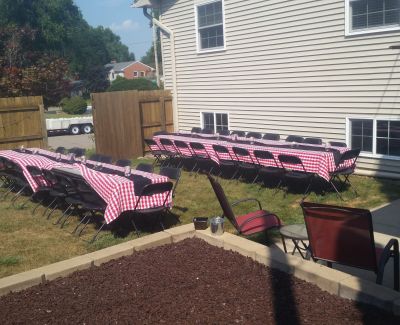 Outdoor backyard party setup with long tables covered in red and white checkered tablecloths and chairs arranged on a grassy area next to a house.