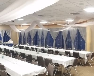 An indoor banquet hall set with long tables covered in white cloths, brown chairs, and decorated with white ceiling swags and blue patterned wall draping behind the head table area.