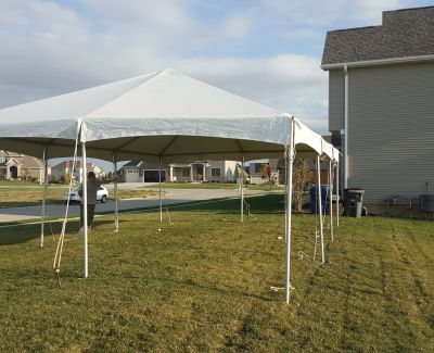 A small white frame tent partially set up on a green residential lawn, secured with ropes and stakes, with houses visible in the background.