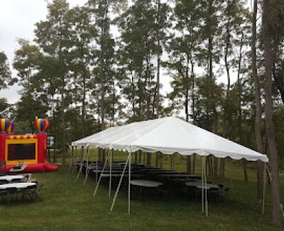 A medium white frame tent set up on a grassy area near tall trees, sheltering several picnic tables arranged in rows. A colorful inflatable bounce house is nearby.