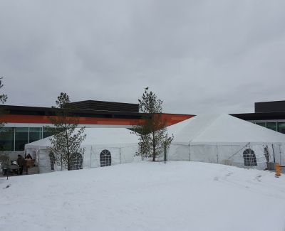 A large white frame tent with windowed sidewalls set up outdoors on snow-covered ground in front of a modern building during winter.