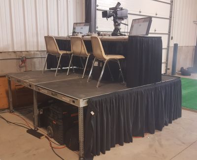 A small portable stage with black skirting, set up with a table, three chairs, laptops, and a broadcast camera on a tripod in front of a warehouse roll-up door.