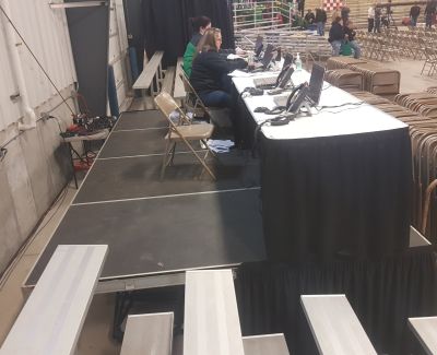 An elevated platform with a black-skirted table, chairs, and laptops, serving as a judging or administrative area overlooking rows of empty bleacher seating below.