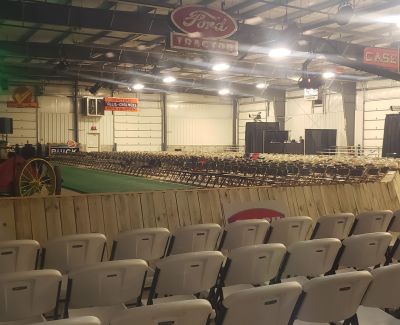 A large arena or exhibition hall filled with rows of white plastic folding chairs facing a central area.