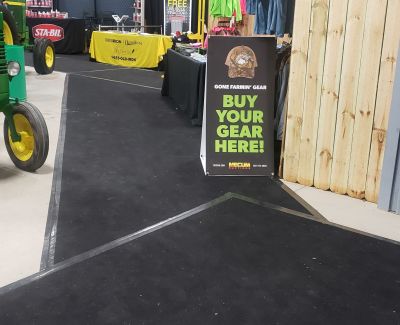 A close-up of a black carpeted walkway at an event, bordered by a green tractor wheel on the left and a wooden partition with a 
