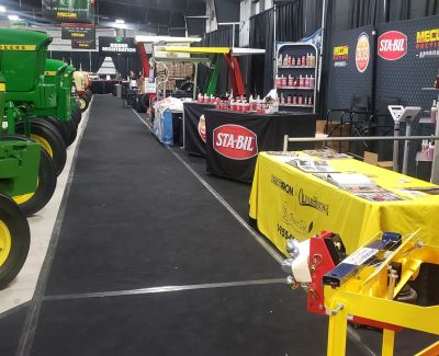 An aisle at an agricultural trade show lined with black carpet, featuring John Deere tractors on the left and vendor booths with displays.