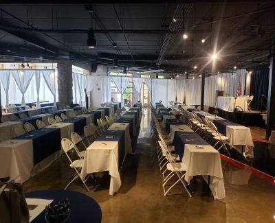 A long view down a conference or banquet room set with multiple rows of tables covered in white cloths and navy blue runners, accompanied by white folding chairs, ready for an event.