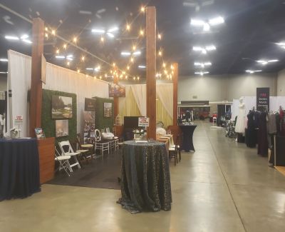 A view down an aisle at an event expo or trade show, featuring various booths, including one prominently decorated with wooden posts, string lights, greenery, and cocktail tables.