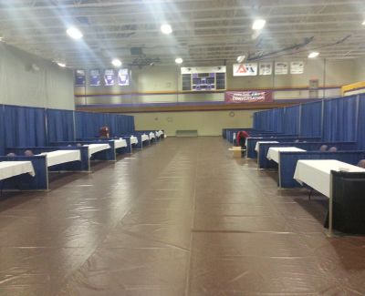 A gymnasium floor, covered with a brown protective layer, is set up for an event with rows of booths defined by blue pipe and drape and white-covered tables lining a central aisle.