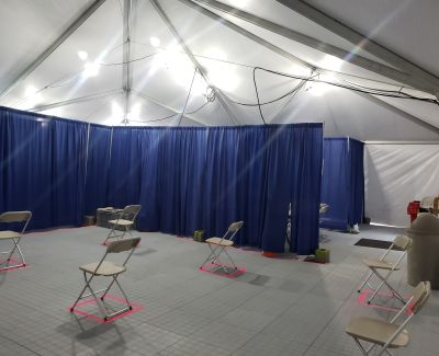 nterior of a large event tent with blue pipe and drape lining the perimeter, showing several folding chairs spaced far apart on a grey modular floor, marked with pink tape squares for social distancing.