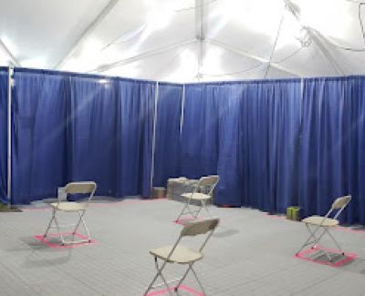 A wider angle inside the event tent showing the blue pipe and drape backdrop and multiple folding chairs positioned individually within pink taped squares.