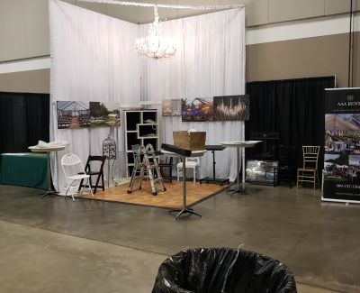 AAA Rent's company's trade show booth featuring white pipe and drape, a chandelier, a small wooden dance floor, shelving units, chairs, tables, and framed pictures showcasing event setups.