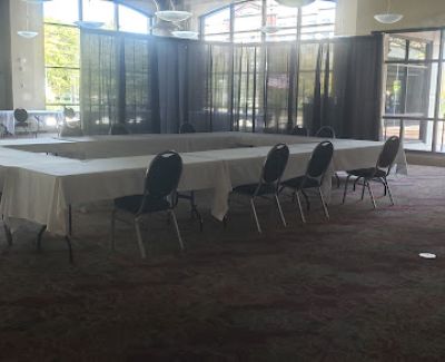 A conference room with tables arranged in a hollow square formation, covered with white tablecloths and surrounded by chairs, set against a backdrop of black pipe and drape near large windows.