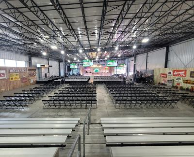 A high-angle view from bleachers overlooking a large venue floor filled with rows of black folding chairs facing a distant stage setup, with additional empty bleachers in the foreground.