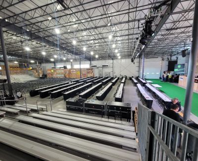 An elevated perspective from bleachers showing rows of long, black-skirted tables arranged on the floor of a large exhibition hall, likely for a trade show or banquet setup.