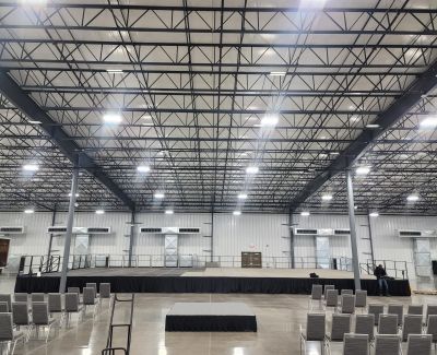 A large, empty event hall with high exposed ceilings and trusses, featuring a raised stage at one end and rows of chairs set up on the floor facing the stage.