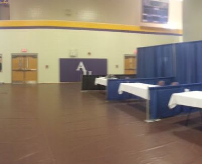 A closer view of an event setup in a gymnasium, showing blue pipe and drape booths, white-covered tables on a brown protective floor.
