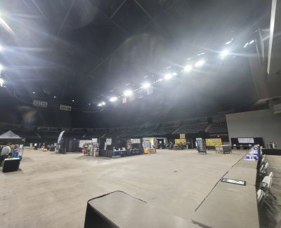 A wide-angle view across a dimly lit arena floor configured for an expo or event, showing scattered vendor booths and equipment under bright overhead spotlights.
