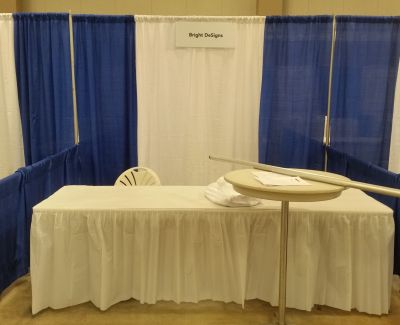 A single trade show booth featuring a white-skirted table, a white chair, a small round pedestal table, and a blue and white pipe and drape backdrop with a 