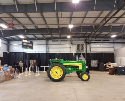 A green and yellow John Deere tractor displayed in the center of a large exhibition hall or warehouse space during an agricultural show or event, with black pipe and drape in the background.