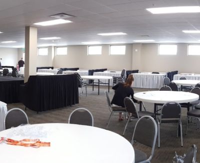 An event space or conference room set up with numerous round tables with white cloths and grey chairs, alongside long rectangular tables covered in black pleated skirts.