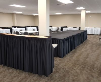 Long rectangular tables covered with black pleated skirts arranged along the walls or in a U-shape within a conference room or event space with carpeted floors and white columns.