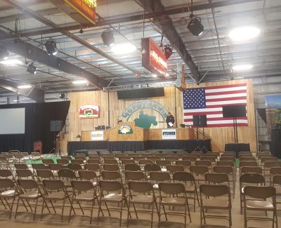 An event hall set up with rows of brown folding chairs facing a wooden stage decorated with an American flag and event branding, prepared for an auction or presentation.