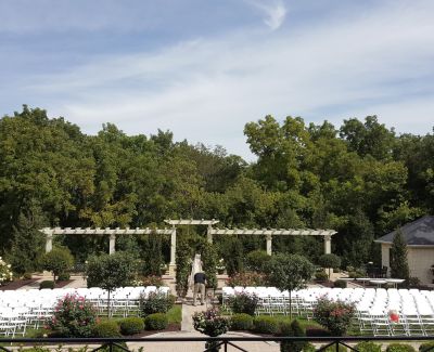 Outdoor garden wedding ceremony setup with rows of white chairs facing a wooden arbor surrounded by greenery.