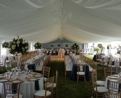Elegant tented wedding reception with chandeliers, navy and gold decor, and floral centerpieces on long banquet tables.