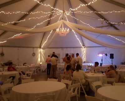 Evening tented wedding with draped fabric ceiling, string lights, chandeliers, and round tables with white chairs.