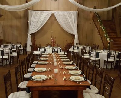 Rustic indoor reception setup with a long wooden banquet table, white drapery backdrop, and wooden staircase.