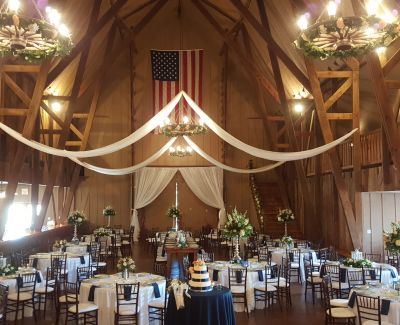 Indoor barn wedding reception with exposed beams, white drapery, floral centerpieces, and a cake table under chandeliers.