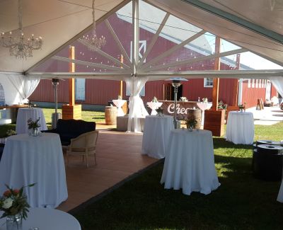 Outdoor tented reception with white drapes, chandeliers, cocktail tables, and a red barn in the background